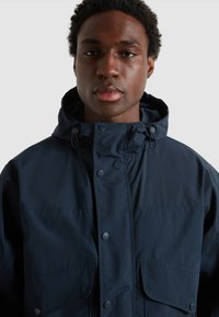 Veste imperméable bleu marine avec capuche, boutons-pression et grandes poches avant, fabriquée dans un tissu lisse et à texture mate.