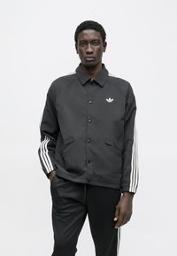 Črna jakna s ovratnikom, sprednjimi gumbi in belimi tremi črtami na rokavih. Ima stranske žepe in vezeno logo.