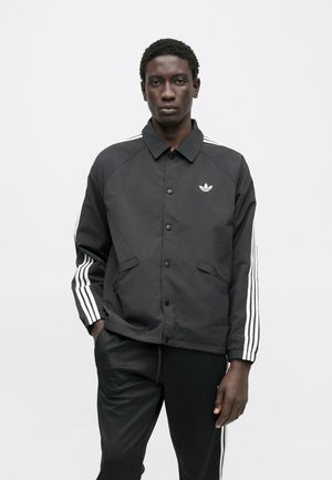 Zwarte jas met een kraag, knopen aan de voorkant en witte drie-strepen accenten op de mouwen. Voorzien van zijzakken en een geborduurd logo.