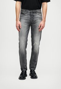 Muškarac nosi izblijedjele crne traperice s rupom sprijeda na desnom bedru, uparen s crnim loafericama i crnom košuljom.