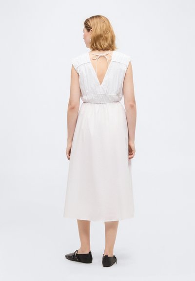 Femme aux cheveux blonds courts portant une robe midi blanche sans manches avec un dos en V profond noué au cou et des chaussures plates noires, debout de dos.