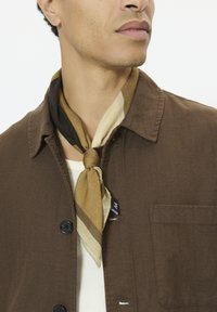 Veste-chemise en coton marron avec des boutons noirs, dotée d'une poche plaquée. Une écharpe rayée multicolore est nouée autour du cou.