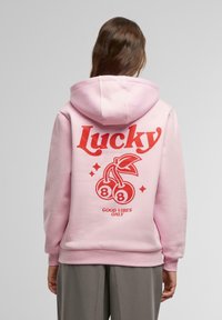 Sudadera rosa con un gran gráfico rojo de cerezas, la palabra "Lucky" y la frase "Good Vibes Only" en la parte posterior. Textura de tela suave.