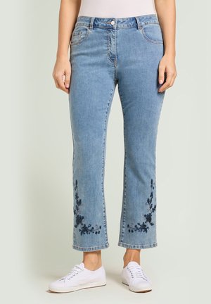 Mujer con vaqueros de mezclilla azul claro con bordado floral oscuro cerca del dobladillo y zapatillas blancas, posando contra un fondo liso.
