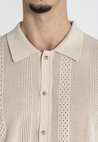 Beige gebreide polo met een gemêleerd ontwerp met verticale strepen, drie knopen en subtiele perforaties voor ventilatie.