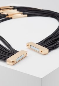 Collar de cordón negro en capas con acentos de metal en tono dorado; los colgantes rectangulares presentan un incrustado blanco y un pequeño detalle de agujero.