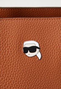 Cuir marron texturé avec une épingle en argent représentant un visage stylisé portant des lunettes de soleil et un bandeau blanc qui y est attaché.