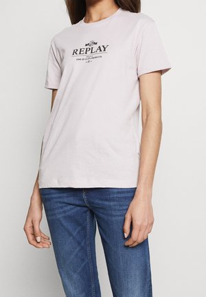 Mujer con camiseta rosa claro de Replay con texto negro y jeans azules, de pie contra un fondo blanco liso.