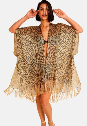 Jeune femme portant un kimono à imprimé tigre avec des franges par-dessus un bikini, posant les mains près de la tête sur un fond uni.