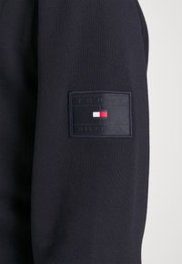 Ciemnoniebieska tkanina rękawa z prostokątną naszywką logo Tommy Hilfiger w kolorach czerwonym, białym i niebieskim.