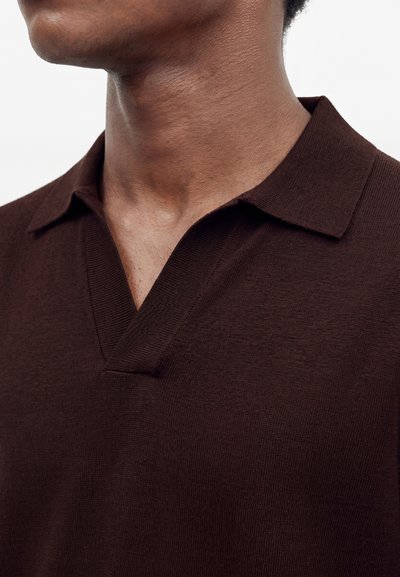 Gros plan d'une personne portant un polo en maille marron foncé avec un col en V ouvert sur un fond clair uni.