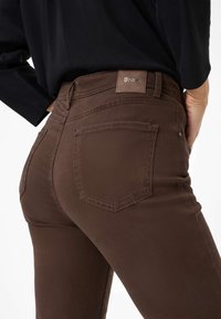 Jeans marroni con una vestibilità slim, realizzati in tessuto morbido. Presentano un'etichetta in rilievo con la scritta "Brax", due tasche posteriori e dettagli di cucitura sottili.