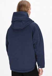 Mörkblå huvjacka med en mjuk textur, förlängd bakre fåll och tryckknappsmuddar. Har en minimalistisk design och dolda dragkedjor.