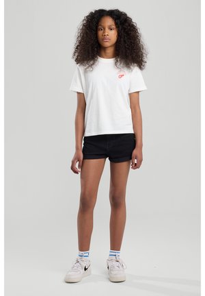T-shirt blanc à manches courtes avec un graphique rouge, associé à un short en denim noir et des sneakers blanches avec des accents noirs et des chaussettes rayées bleues.