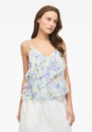 Žena s dlhými hnedými vlasmi, oblečená v pastelovom kvetinovom volánovom tielku bez rukávov a bielej sukni, stojaca pred jednofarebným pozadím.