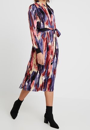 Kvinde iført en langærmet midikjole med akvarelmønster i rød, lilla og creme, med et sort bælte i taljen og sorte ankelstøvler.