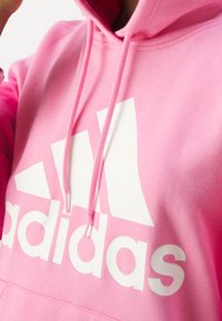 Rosa bomullshuvtröja med en vit Adidas-logotyp, snoddar och en mjuk, texturerad yta. Närbilden framhäver logotypen och tyget.