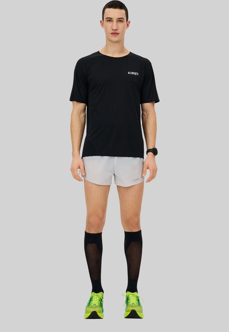 Schwarzes Sporthemd mit kurzen Ärmeln, hellgraue Shorts, kniehohe schwarze Kompressionssocken und grüne Schuhe; minimalistisches Design, atmungsaktiver Stoff.