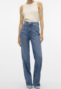 Top senza maniche di colore chiaro abbinato a jeans blu a vita alta con dettagli decorativi di borchie vicino all'orlo.