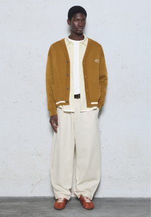 Homme portant un cardigan marron sur une chemise rayée et un tee-shirt blanc, pantalon blanc ample et baskets marron, debout contre un mur clair uni.