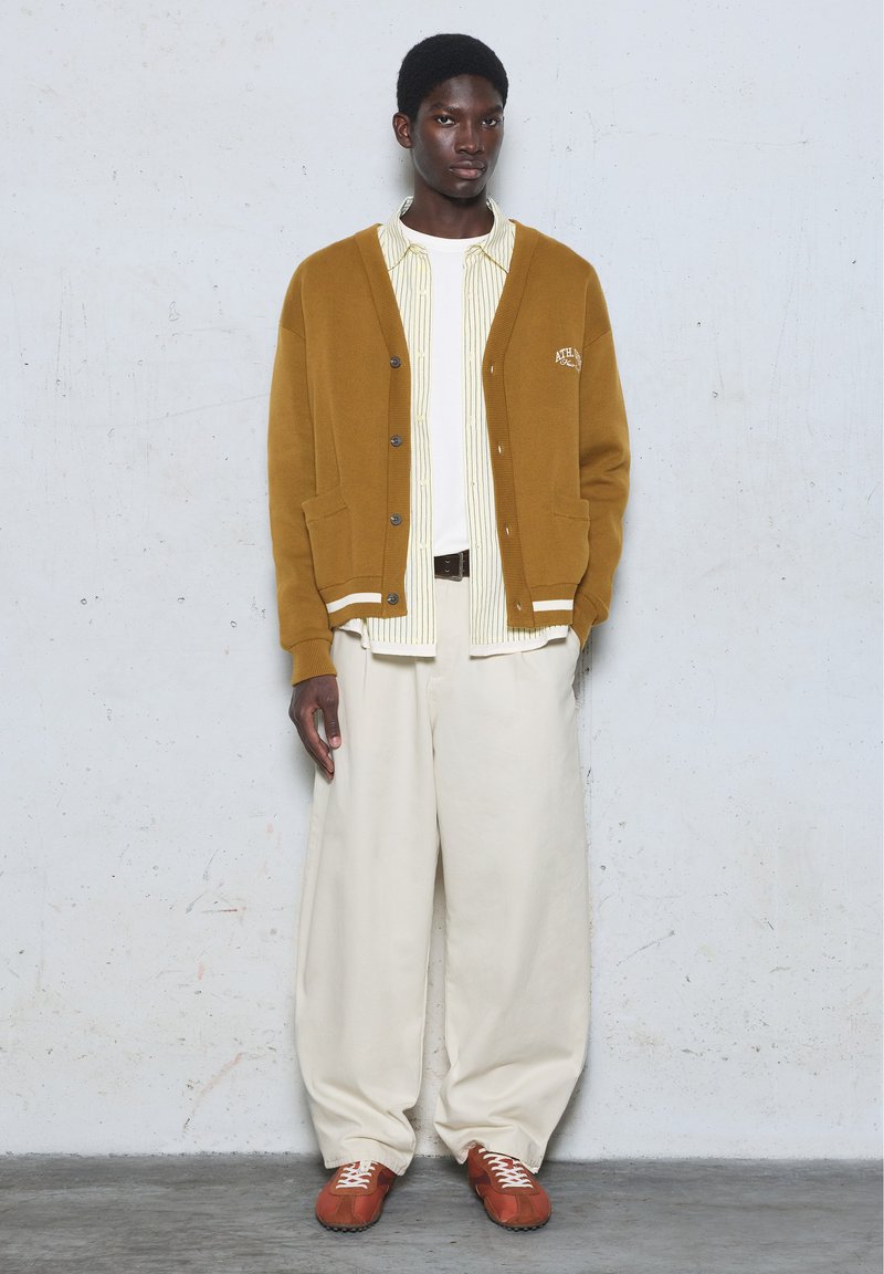 Homme portant un cardigan marron sur une chemise rayée et un tee-shirt blanc, pantalon blanc ample et baskets marron, debout contre un mur clair uni.