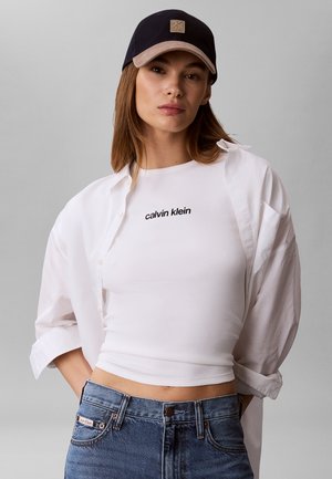 Kvinde iført sort kasket, hvid Calvin Klein crop top, oplukket hvid skjorte og blå jeans mod ensfarvet grå baggrund.
