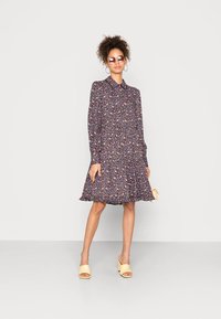 Vestido de manga longa, com colarinho, apresenta um padrão floral escuro, com bainha franzida e formato esponjoso. Combinado com sapatos de salto aberto amarelos.