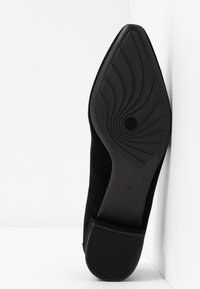 Semelle de chaussure noire pointue avec motif de bande de roulement en spirale et talon rectangulaire, appuyée verticalement contre un mur d’angle blanc.