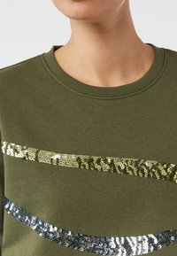 Oliwkowa zielona bluza z okrągłym dekoltem. Posiada poziome linie złotych i srebrnych cekinów na wysokości klatki piersiowej, tworzące teksturowany wzór.