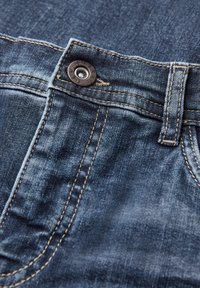 Vaqueros de mezclilla en azul oscuro con una textura suave, que presentan costuras doradas en contraste y un botón de metal en la cintura.
