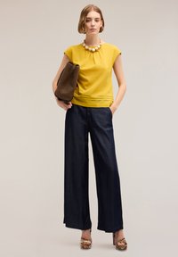Geel top met korte mouwen en geplooide halslijn, gecombineerd met donkerdenim wijde broek. Accessoires bestaan uit een gestructureerde bruine tas en geprinte schoenen.