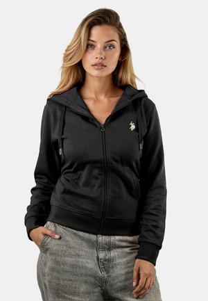 Schwarze Zip-Hoodie aus weichem Material mit Kapuze, Kordeln und einem kleinen goldenen Emblem auf der linken Brust. Kombiniert mit grauen Hosen.