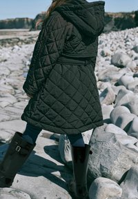 Enfant portant un manteau matelassé noir et des bottes noires marchant sur de gros rochers lisses près d'une côte rocheuse par une journée claire.