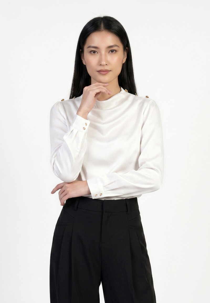 Jeune femme aux cheveux noirs et lisses portant un chemisier blanc et un pantalon noir, debout avec une main sous le menton et l’autre croisée.