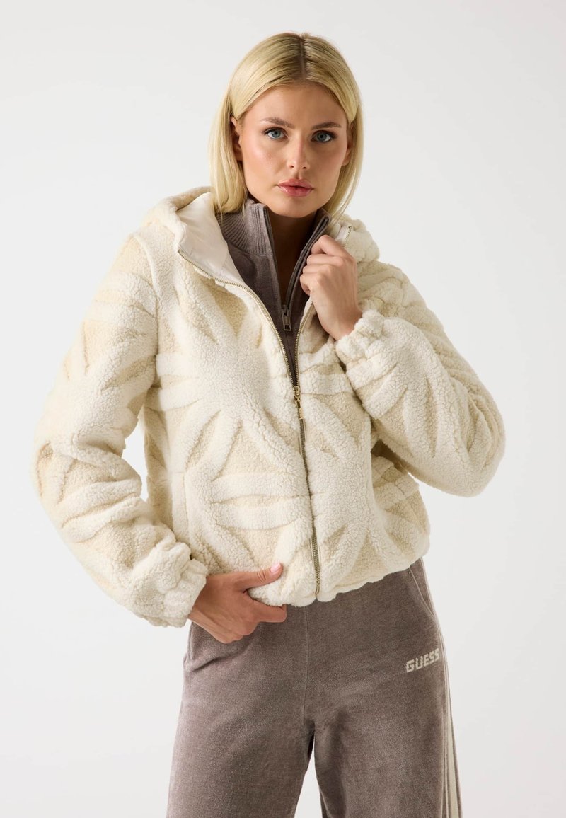Giacca in finta pelliccia con cappuccio di colore crema, con motivo a foglia texture, chiusura con zip e polsini elastici. Indossata sopra un top zip beige chiaro e pantaloni coordinati.