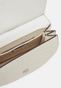 Texturerad krämfärgad handväska med en slät, vit klaff, som har flera innerfack och en metallknäppning.