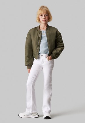 Veste bomber vert olive, haut en denim clair, jeans blancs évasés, baskets blanches, pose debout, arrière-plan gris neutre.