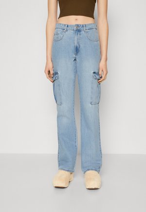 Person iført lyseblå cargo jeans med lige ben og klaplommer samt beige, plyssede hjemmesko, mod en ensfarvet hvid baggrund.