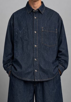 Uomo che indossa una camicia di jeans blu scuro con bottoni, tasca frontale e un sottile motivo ricamato a "XX", abbinata a pantaloni di jeans dello stesso colore.