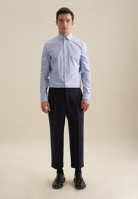 Camicia button-up azzurro chiaro con colletto classico, abbinata a pantaloni eleganti scuri e scarpe in pelle nere, su uno sfondo neutro.