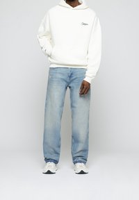 Sweat à capuche blanc avec poche avant et logo discret, associé à un jean droit bleu clair et des baskets blanches. Tenue décontractée.