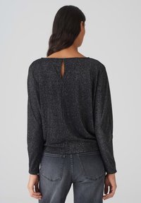 Schwarzes, langärmliges Oberteil mit schimmernder Textur, das ein Schlüsselloch-Detail auf der Rückseite und eine lockere Passform aufweist, kombiniert mit grauen Jeans.