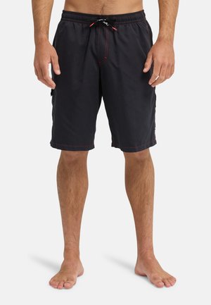 Mężczyzna w czarnych, sięgających do kolan szortach kąpielowych z czerwonymi przeszyciami i sznurkiem, stoi boso na białym tle.