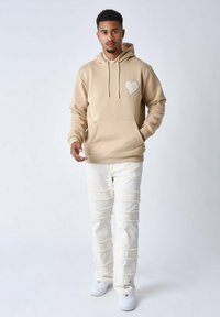 Sudadera beige con un detalle en forma de corazón, cordones y bolsillo frontal, combinada con jeans blancos desgastados que tienen tiras deshilachadas horizontales y zapatos blancos.