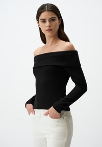 Zwarte off-the-shoulder top van ribstof met lange mouwen en een aansluitend ontwerp. Gecombineerd met lichtgekleurde jeans.