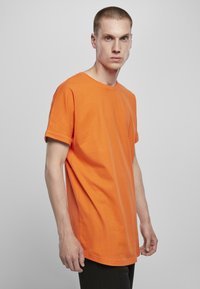 Tricou din bumbac portocalie cu mâneci răsucite, decolteu rotund, croială lejeră și textură fină, purtat peste pantaloni negri pe un fundal neutru.