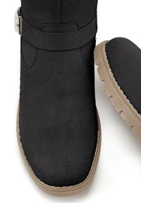 Schwarze Wildleder-Stiefeletten mit strukturiertem Finish, ausgestattet mit einem grauen Riemen und einer hellbraunen Gummisohle. Rundliche Zehenform mit sichtbaren Nahtdetails.