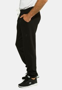 Schwarze Jogginghose mit einem Kordelzugbund, Seitentaschen und einem dezent geprägten Logo am linken Bein. Das Material sieht weich und elastisch aus.