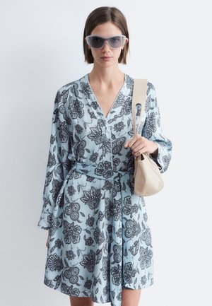 Vrouw die een lichtblauwe gebloemde jurk draagt met een bijpassende riem, oversized zonnebril en een beige handtas over haar schouder hangt.