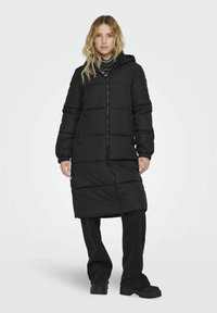 Manteau d'hiver noir long avec sections matelassées, fermeture éclair à l'avant et capuche. Éléments des poignets en élastique et deux poches à l'avant. Tissu texturé.
